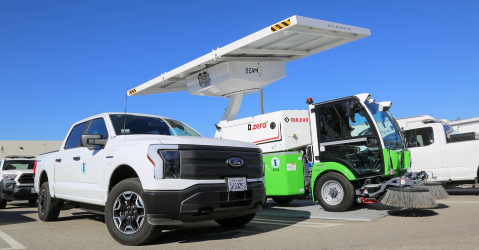 A white electric truck and green and white street sweeper are parked under a solar-powered EV charger.